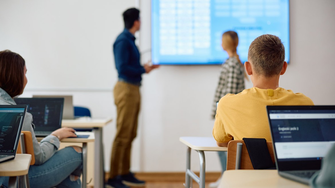 Um professor sorri enquanto usa um tablet para planejar uma aula, com ícones de ideias e criatividade ao redor, simbolizando o uso da IA para educadores.